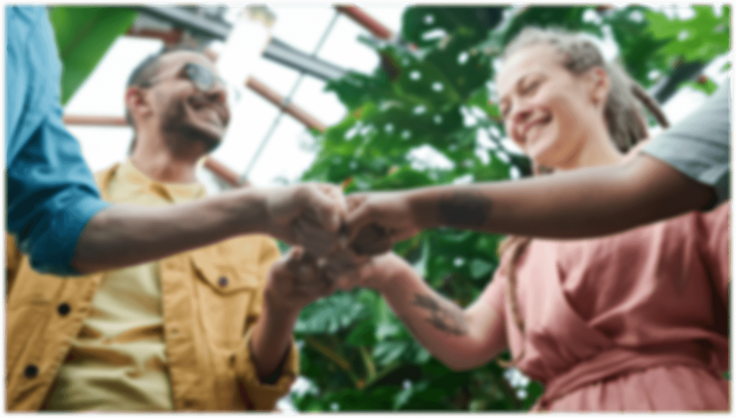 A diverse group of people holding hands in a lush greenhouse, surrounded by vibrant green plants and colorful flowers.